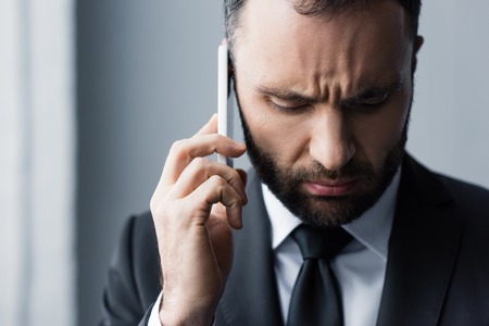 Serious bearded man in black suit looking down while using smartphoneの写真素材