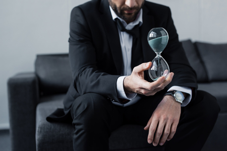Partial view of sitting businessman in black suit holding hourglassの写真素材