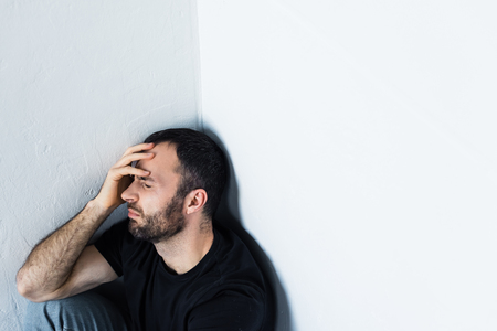 Depressed man sitting in corner with closed eyes and holding hand on foreheadの写真素材