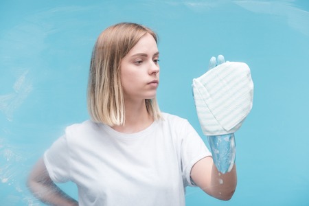 Young woman in rubber glove cleaning glass with rag on blue backgroundの写真素材