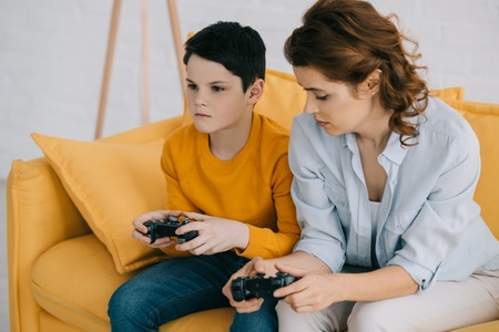 attentive boy playing video game with joystick while sitting on sofa near pensive motherの写真素材