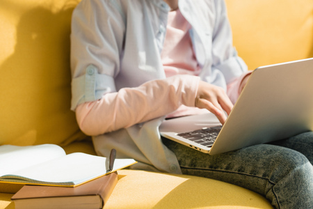 partial view of child using laptop while sitting on yellow sofa near book and copy bookの写真素材