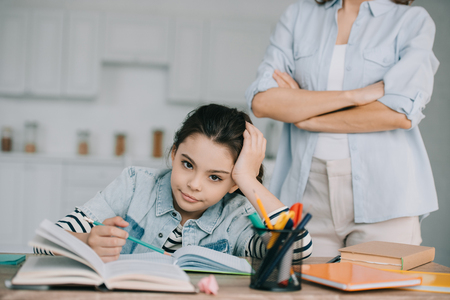 partial view of woman standing with crossed arms behind bored daughter doing homeworkの写真素材