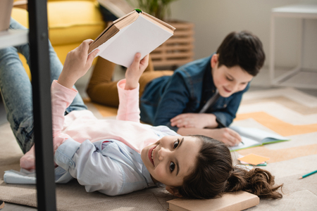 selective focus of cheerful child lying on floor with book near brother writing in notebookの写真素材