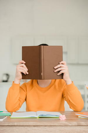schoolboy hiding face behind book while sitting at desk and doing homeworkの写真素材