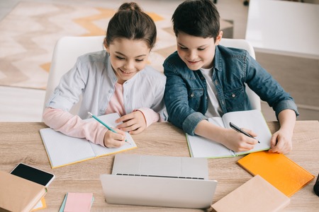 smiling brother and sister writing in notebooks and using laptop togetherの写真素材