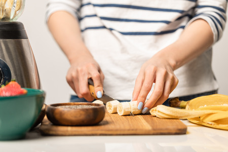 cropped view of woman cutting organic and ripe bananas on chopping board while standing on whiteの写真素材