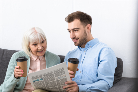 smiling man and surprised senior mother holding coffee and reading newspaper at homeの写真素材