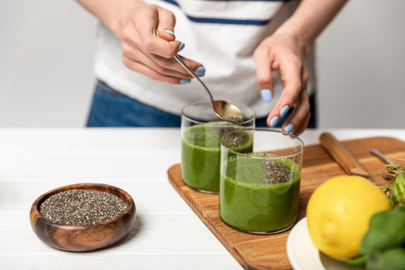 cropped view of woman holding spoon with chia seeds near glasses of green smoothie on greyの写真素材