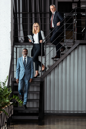 cheerful blonde businesswoman walking on stairs near multicultural businessmenの写真素材