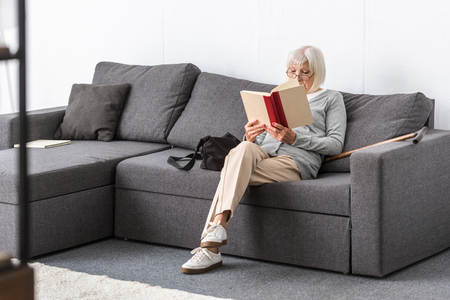 senior woman in glasses sitting on sofa and reading book in living roomの写真素材