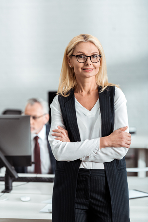 selective focus of cheerful blonde businesswoman in glasses standing with crossed arms near coworkerの写真素材