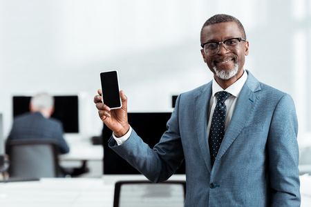 selective focus of happy african american businessman holding smartphone with blank screen in officeの写真素材