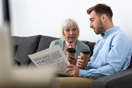 man and senior mother holding coffee and reading newspaper at homeの写真素材