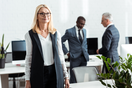 selective focus of cheerful blonde woman standing in office near multicultural businessmenの写真素材