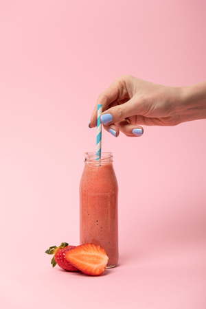 cropped view of woman holding straw in glass bottle with tasty smoothie near ripe strawberries on pinkの写真素材