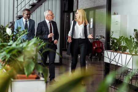selective focus of businesswoman gesturing while walking with multicultural partners in officeの写真素材