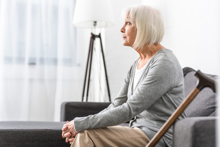 pensive senior woman with wooden cane sitting on sofa in living roomの写真素材