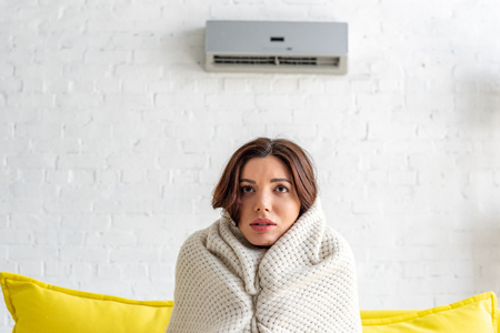 frozen young woman warming under blanket while sitting on sofa under air conditioner at homeの写真素材