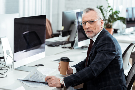 handsome mature businessman holding newspaper and paper cup in officeの写真素材