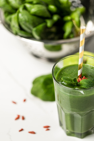 selective focus of tasty smoothie with goji berries and mint in glass with straw near spinach leaves on whiteの写真素材