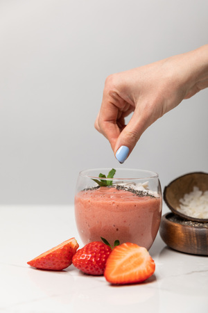 cropped view of woman adding coconut flakes in tasty smoothie near red strawberries on greyの写真素材