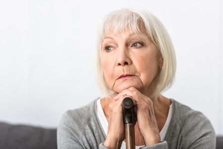 pensive senior woman with wooden cane looking away at homeの写真素材