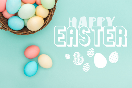 Top view of multicolored painted eggs in wicker basket with happy Easter lettering on blue...