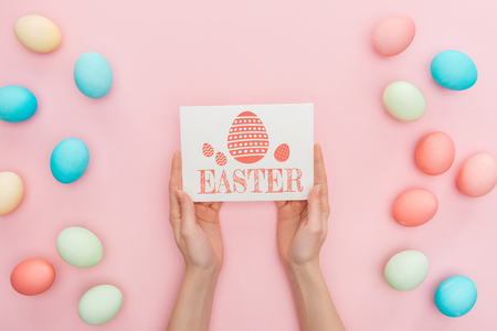 Cropped view of woman holding greeting card with Easter lettering near multicolored painted chicken eggs on pink backgroundの写真素材
