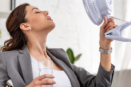 Beautiful businesswoman holding electric fan and glass of water with closed eyes while suffering from heat in officeの写真素材