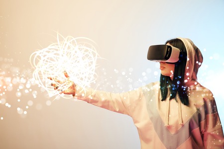 Young woman in virtual reality headset pointing with hand at glowing cyber illustration on beige and blue backgroundの写真素材