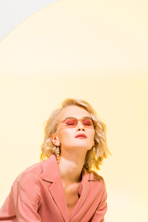 Beautiful stylish blonde girl in coral jacket posing on yellow background with copy spaceの写真素材