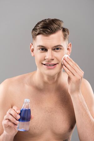 Front view of smiling naked young man holding lotion bottle and wiping face with cotton pad isolated on grey backgroundの写真素材