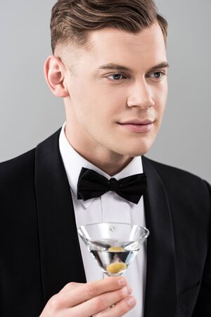 Smiling young man in formal wear with bow tie holding glass of cocktail isolated on grey background