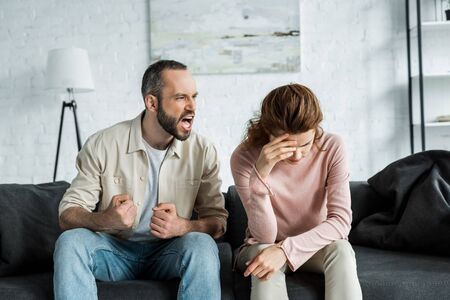 Angry man sitting on sofa and screaming at upset woman