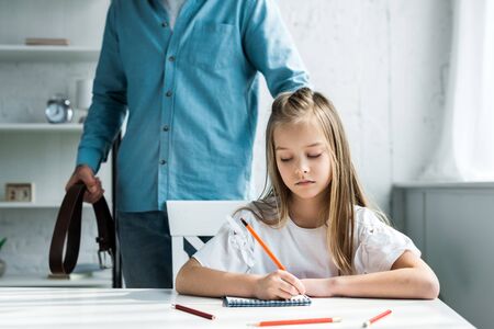 Cropped view of father holding belt and standing near cute kid holding pencil near notebookの写真素材