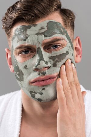 Displeased young man with clay mask touching face isolated on grey backgroundの写真素材