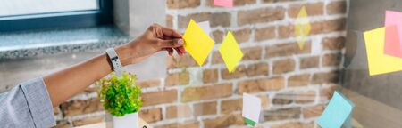 Panoramic shot of African American woman putting Sticky Notes on glass window in loft office
