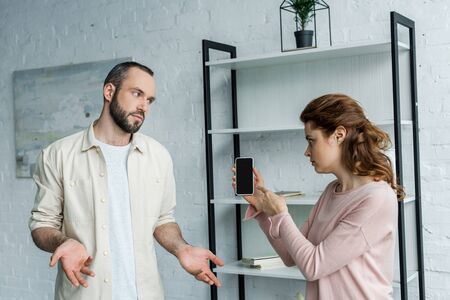 Offended woman pointing with finger at smartphone with blank screen near man at home