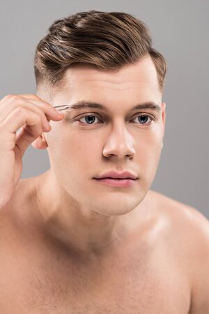 Focused young man using eyebrow tweezers isolated on grey background