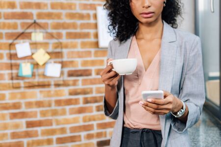 Cropped view of African American casual businesswoman with coffee cup using smartphoneの写真素材