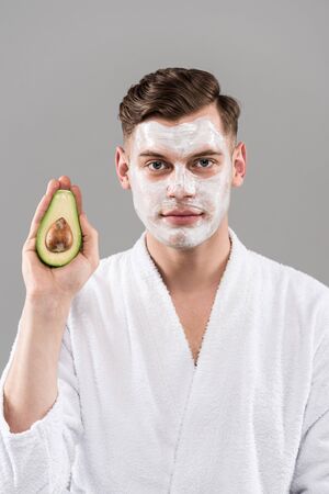 Front view of man in bathrobe with cream on face holding cut avocado isolated on grey backgroundの写真素材