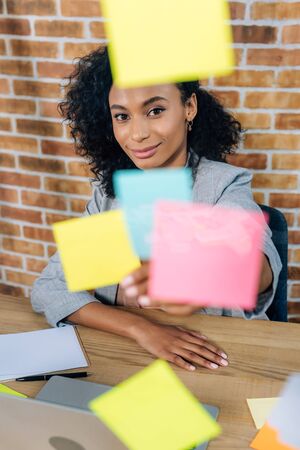 Beautiful African American casual businesswoman sitting at desk in front of glass window with sticky...
