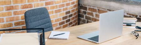 Panoramic shot of wooden desk with laptop, notebook and pen in loft office
