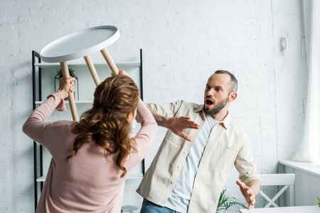 Selective focus of surprised man looking at angry holding chair while threatening at home