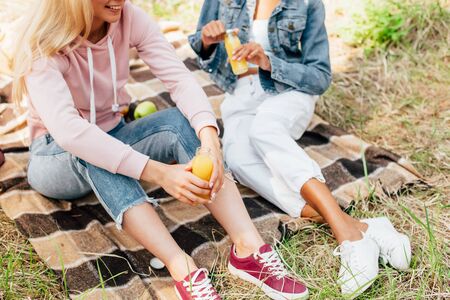 Cropped view of two multiethnic girls sitting on plaid blanket and holding bottles of orange juiceの写真素材