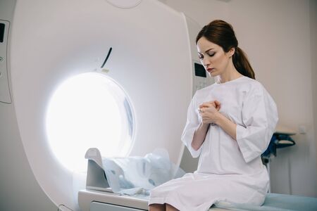 Beautiful woman praying with closed eyes while sitting on mri scanner bed in hospitalの写真素材