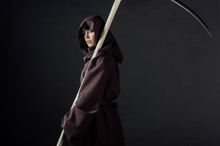 Woman in death costume holding scythe and looking at camera isolated on black backgroundの写真素材