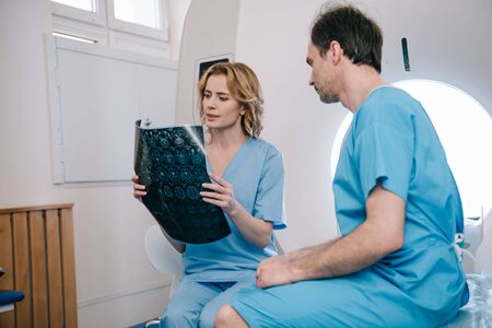Attentive radiographer looking at x-ray diagnosis near patient sitting on ct scanner bed