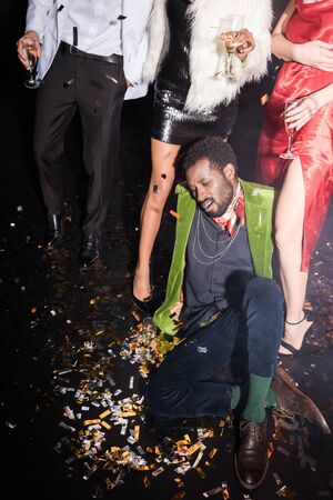 Handsome and drunk African American man lying on floor with shiny confetti near friends on black...